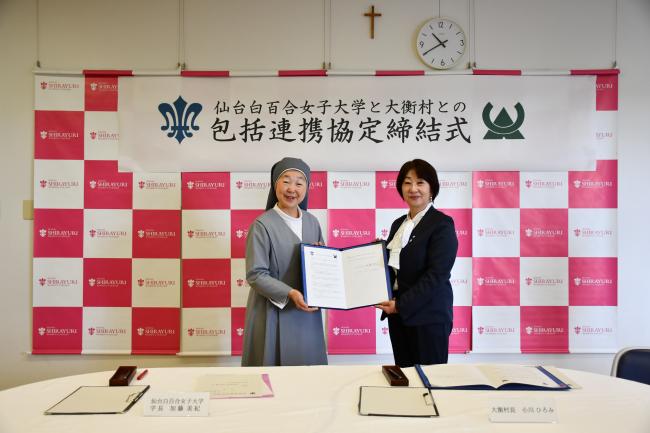 白百合女子大学と大衡村との包括的連携協定締結式と書かれた幕の前で締結書を持つ二人の女性の写真