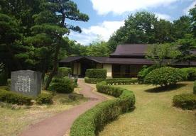 青空の下、木々が生える庭園に佇む日本家屋の写真