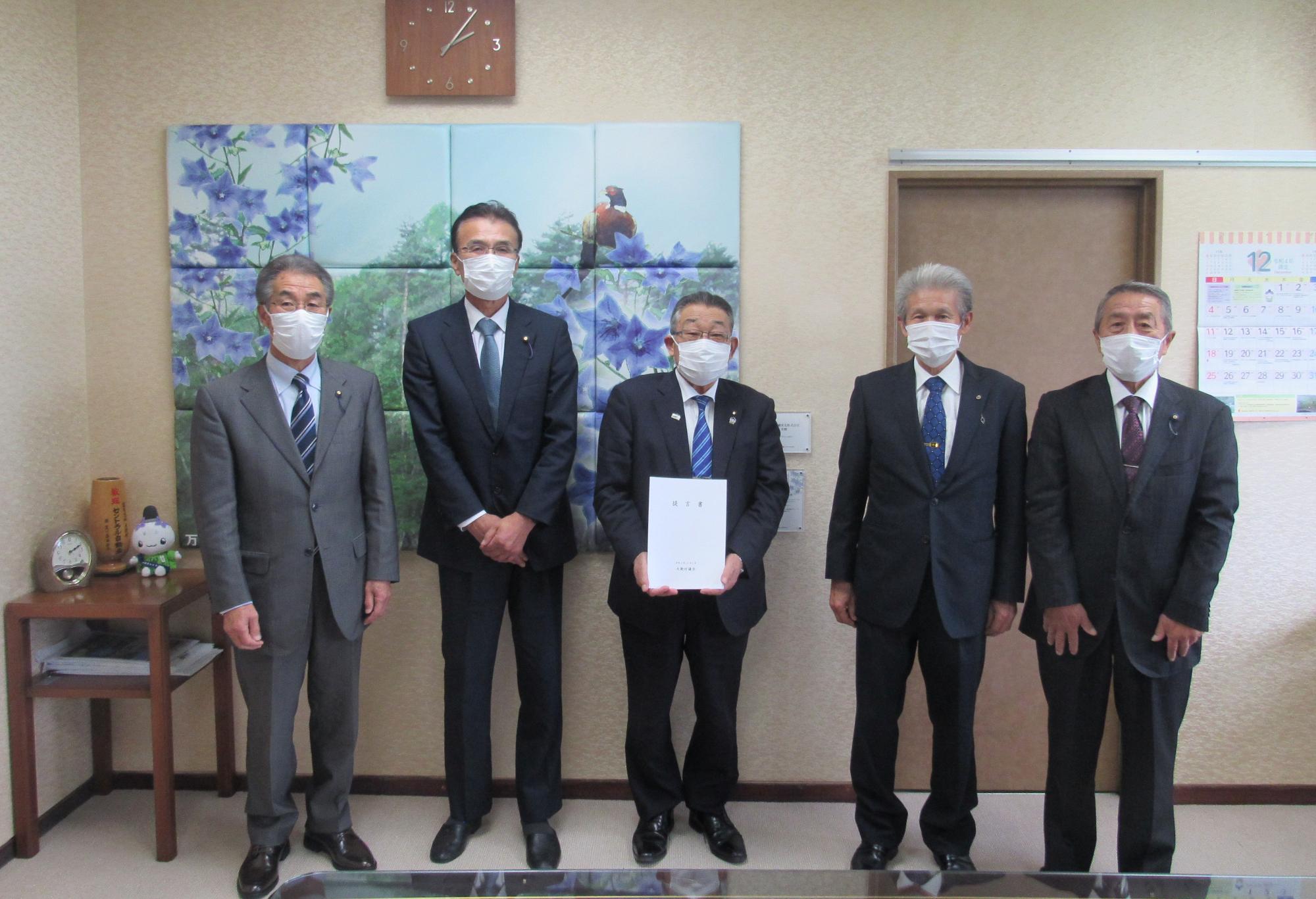 スーツを着た男性に囲まれている真ん中に書状を持った男性の写真