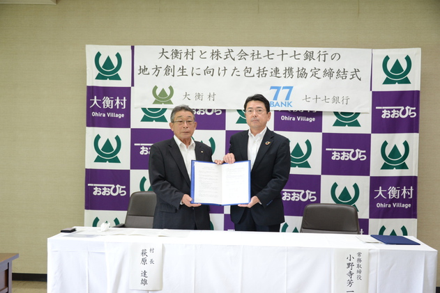 大衡村と株式会社七十七銀行の地方創生に向けた包括連携協定締結式と書かれた幕の前に書類をもって立つ2人の男性の写真