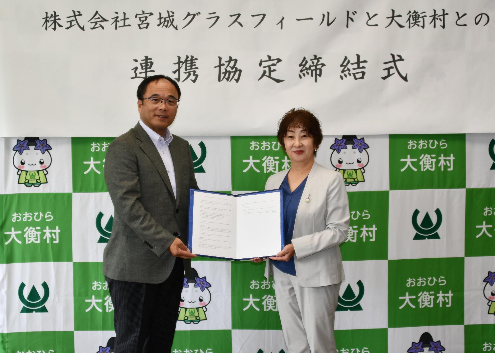 株式会社グラスフィールドと大衡村との連携協定締結式と書かれた幕の前で締結書を持つ男性と女性の写真